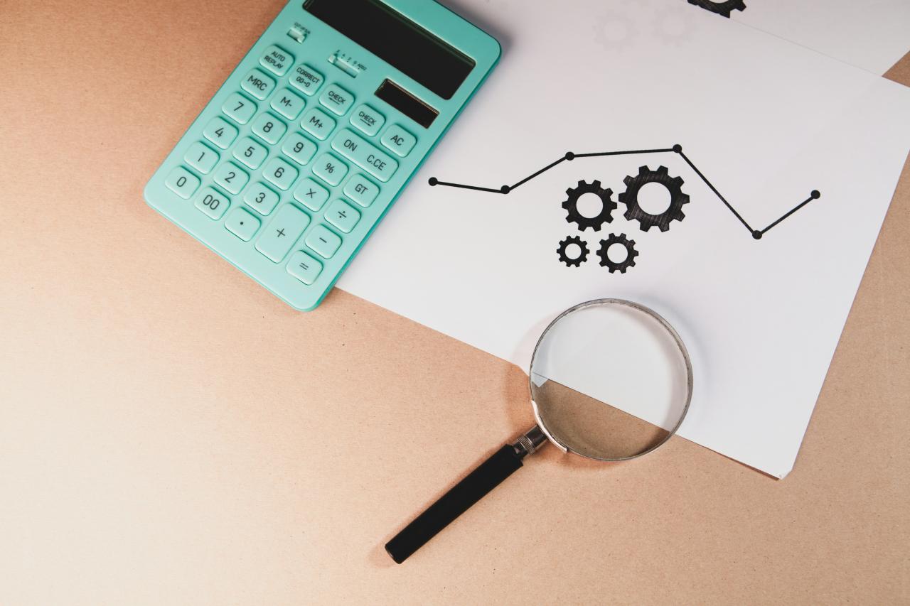 Kalkulator, kaca pembesar, dan bagan dengan roda gigi di atas kertas.