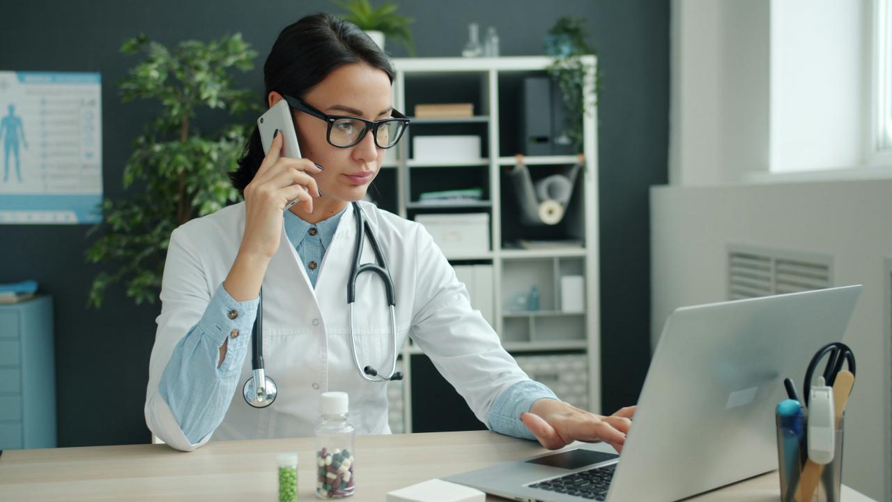 Dokter di telepon, bekerja di laptop di kantor.